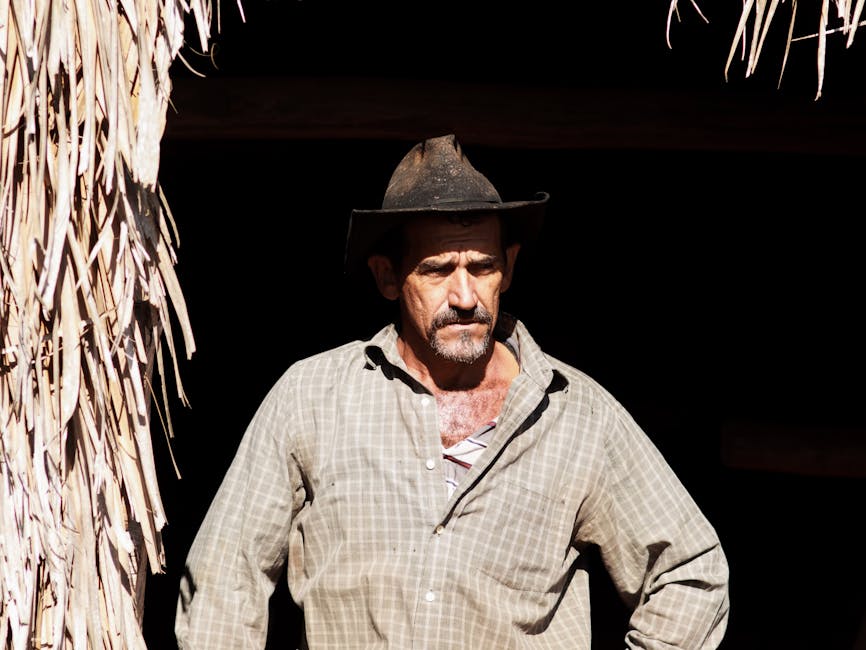 A rugged man stands in bright sunlight, wearing a wide-brimmed hat and plaid shirt.
